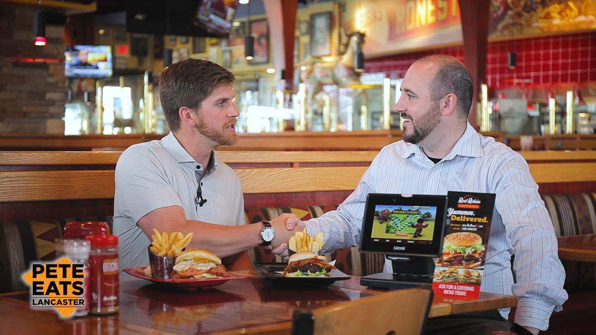 LancasterOnline host Pete Andrelczyk and general manager Cassidy Bailey shake hands, seated at a restaurant table with burger plates and tabletop tablet in front of them