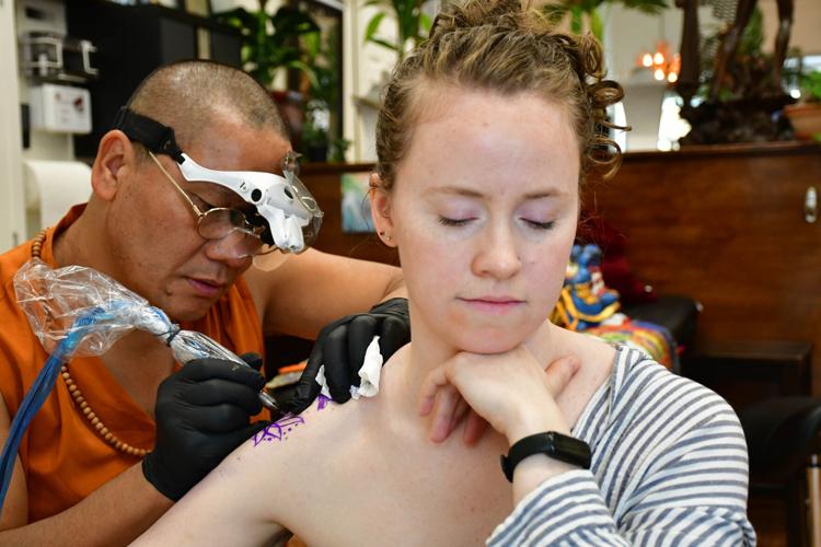 A woman gets a tattoo from a tattoo artist and monk