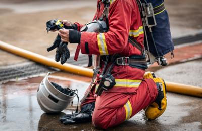 A firefighter wearing a detailed red protective suit with yellow reflective strips kneels on wet pavement during the daytime