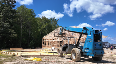 A blue forklift in front of an unfinished home under construction, next to a forest.
