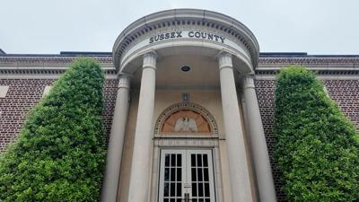 A low angle view of a tall brick building with four pillars, two trees on either side of the entrance, with the words "Sussex County" at the top.