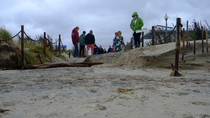 A group of people in coats stand at the beach entrance, where there's a foot drop due to erosion. A rope fence and dune grass surround the entrance on both sides.