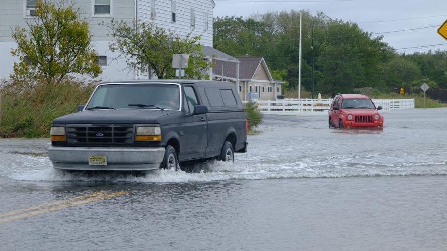 A gray truck is followed by a red Jeep as both drive through water on the street, making splashes around their wheels.