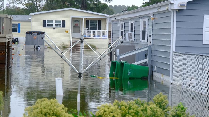 Two green garbage cans lay on their sides with lids open next to a gray house surrounded by floodwaters. A clothesline is in the foreground.
