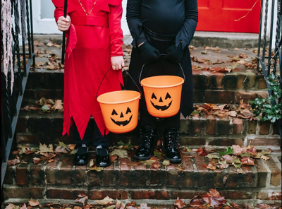Two children standing on steps in a red and black costume while holding orange jack o'lantern candy buckets.