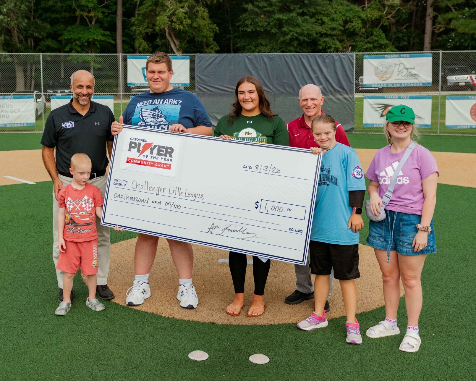 Athletes and parents stand on a sports field holding a large check.