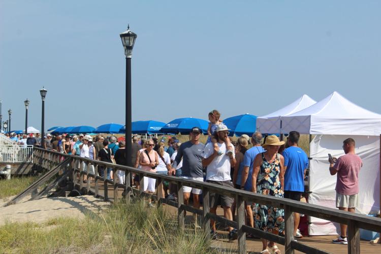 people walk on a boardwalk, a large crowd of people, blue and white tents line the boardwalk on a sunny day