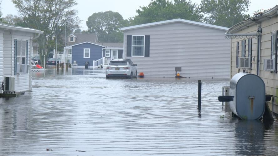 A group of manufactured homes is surrounded by water, showing no grass or streets. A car is parked up close to one house, also sitting in water.