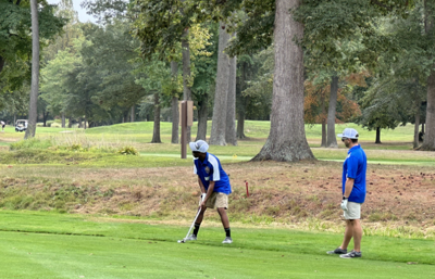 Two people stand on a golf course, one holds a golf club  while another looks on.