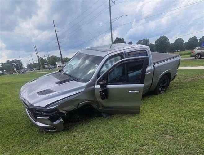 A gray pickup truck slumped over in a grassy field, with the driver's door open and the front left-side wheel missing.