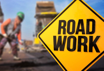 A yellow diamond shaped sign with the words Road Work in all capital letters. In the background is a person doing doing road work.