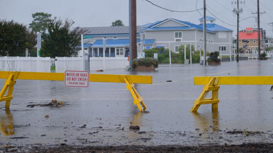 Yellow temporary fences block a road that is covered in water, mulch, and dirt. A sign on one fence says water on road, do not enter, violators fined.