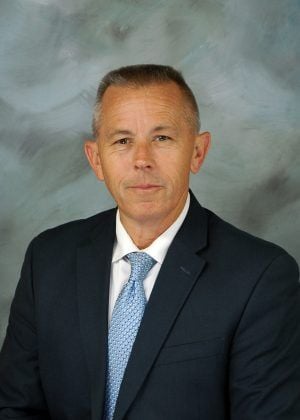 Man posing for headshot with gray background wearing black suit and light blue tie