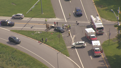 A white car and a black car sit amongst debris at an intersection, surrounded by emergency services.