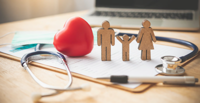 Wooden figurines of a family on a clipboard next to a red heart and surrounded by a doctors stethoscope.