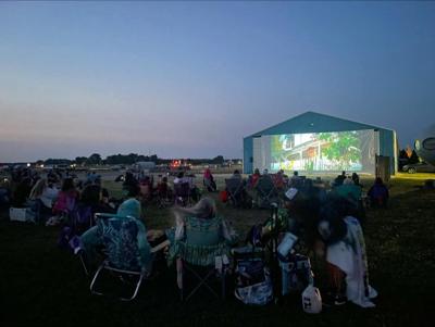 People sitting on folding chairs watching an outdoor movie on a big screen.