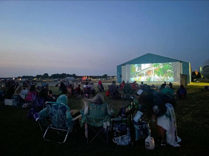 People sitting on folding chairs watching an outdoor movie on a big screen.