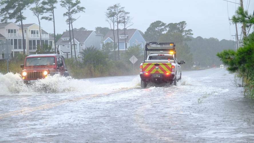 A work truck and a red Jeep drive through deep water on a road.