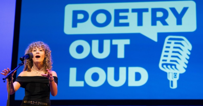 a woman stands in front of a mic with a sign that read 'poetry out loud' behind her in blue and white writing. 