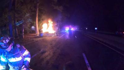 Flames shooting upward from a car that crash into a tree on the left side of the road. A firefighter is the lower left side.