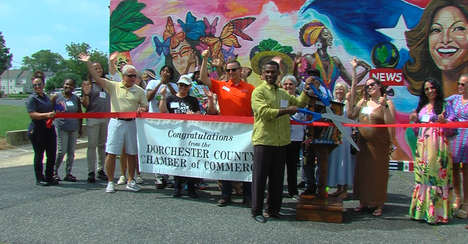 A group of people stand in front of a mural with a ribbon and sign, about to cut the ribbon with large scissors.