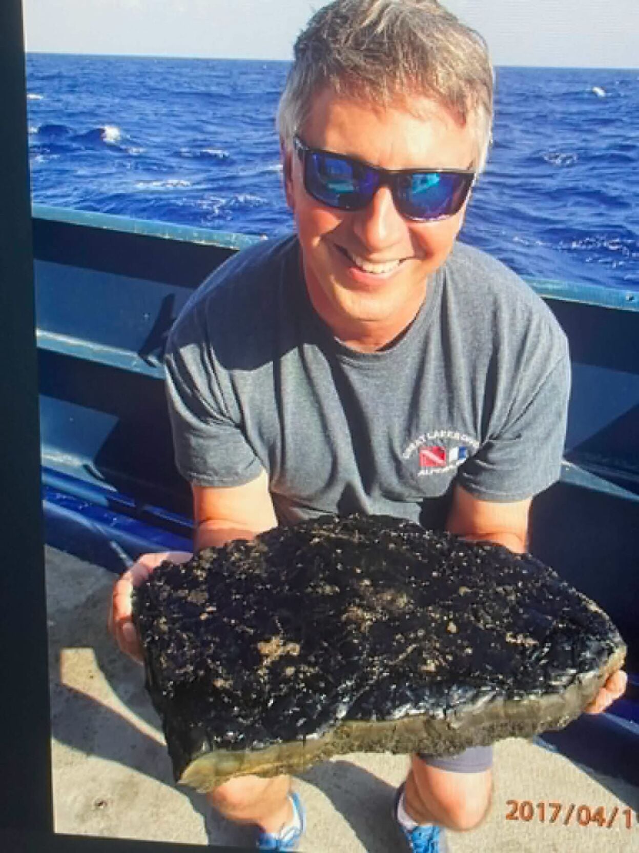 Geppert is squatting on a boat in the water, holding a large rock.