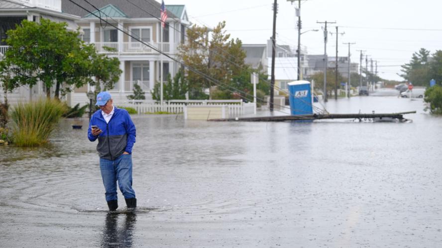 A man wearing a blue and gray jacket walks through water on a residential street. A fallen power pole and portapotty are in the background.