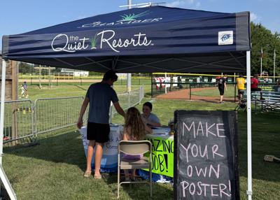 Three people sit under a tent alongside a softball field with a sign that says, "Make your own poster!"