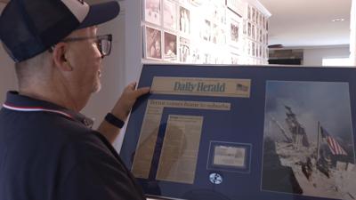 Joe Dittmar, wearing a 9/11 hat looks at framed reminder of the terrorist attack. On the left is a newspaper clipping, in the center is his south tower World Trade Center visitor's badge from that day. On the right is a picture of the American flag being raised in the rubble.