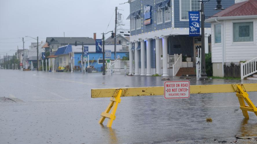A yellow temporary fence blocks a road that is covered in water. A sign on the fence says water on road, do not enter, violators fined.