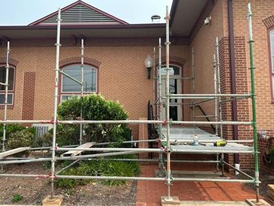 A brick building with windows and a masonry sidewalk. Scaffolding and construction equipment sit in front of building as an ADA ramp is constructed.
