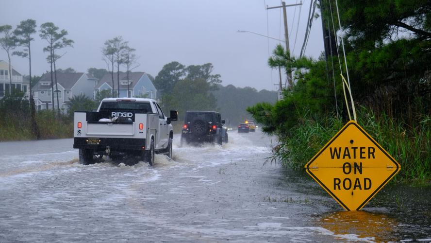 A white truck follows another vehicle through a flooded street in front of a sign that says water on road.