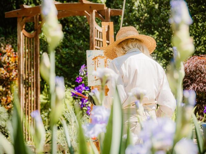 An image showing a woman painting in a sunlit garden surrounded by flowers and greenery.