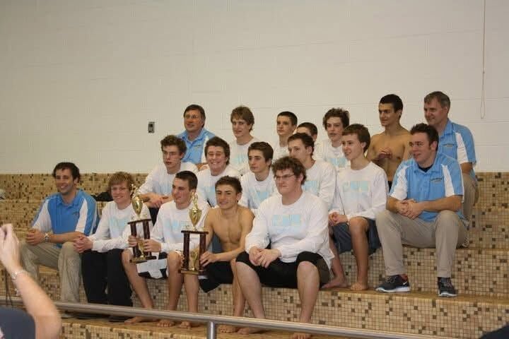 Bill Geppert sits with a swim team on bleachers, holding trophies.