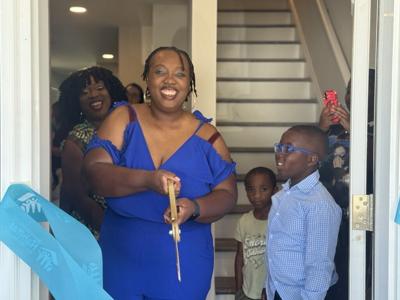 A mother stands at an open door in a blue dress standing among a child in a blue shirt looking up at her. in the background is a smaller child standing by the steps in the entryway of the home. 