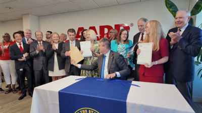 A large crowd of people gather around a table with the governor, holding papers.