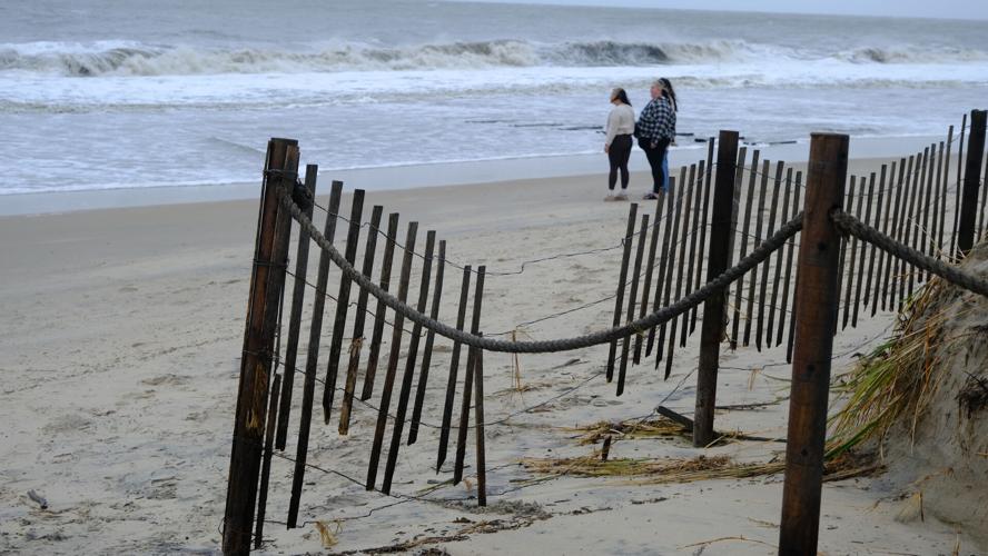 A wooden fence on a beach missing multiple vertical slats with waves in the background.