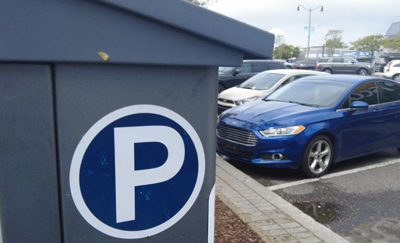 A parking kiosk with a large white letter P in a blue circle on the side. Several cars are parked head-first to the right of the kiosk.