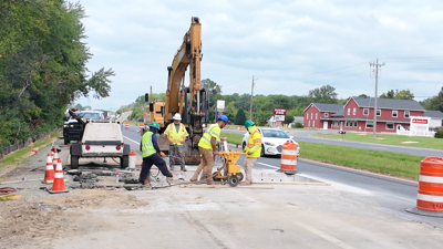 Road crews work cut the asphalt on route one while standing in front of an excavator.