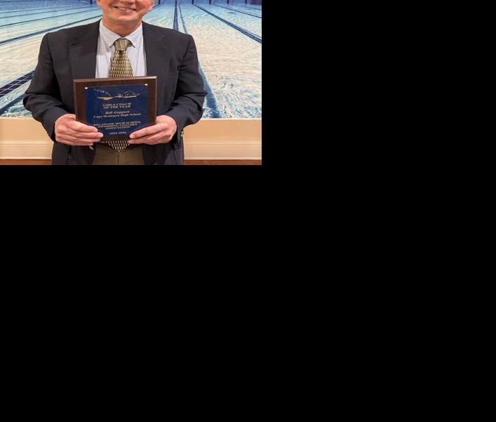 Geppert stands in a suit in front of a wall with a picture of under the water in a lap pool while holding a plaque. 
