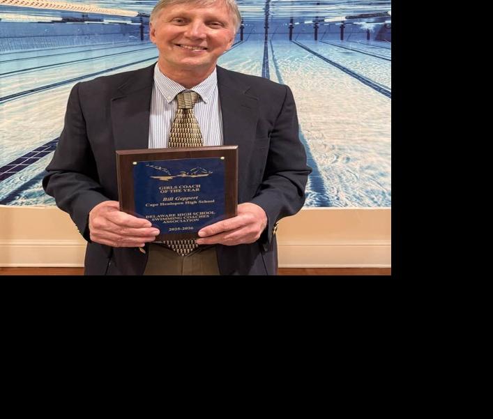 Geppert stands in a suit in front of a wall with a picture of under the water in a lap pool while holding a plaque. 