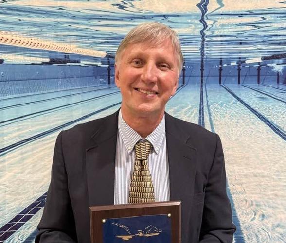 Geppert stands in a suit in front of a wall with a picture of under the water in a lap pool while holding a plaque.