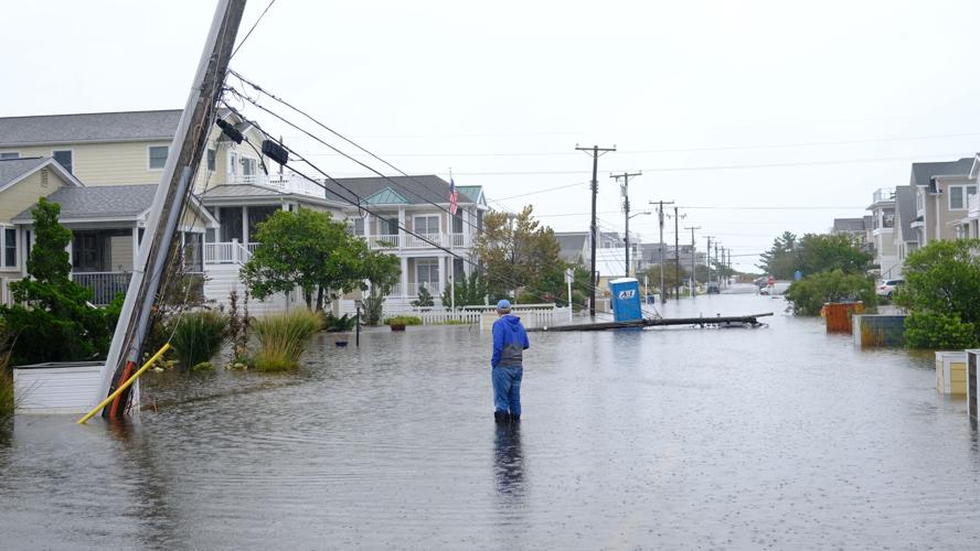 A man wearing a blue jacket stands in the middle of a flooded street looking at a fallen power pole.