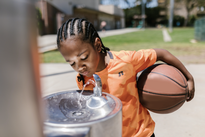 A boy drinks from a water fountain in a basketball school park