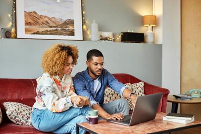 Young man talking with girlfriend while pointing on laptop at coffee table. Afro woman is sitting with boyfriend on sofa. They are in living room.