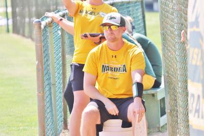 A softball coach wearing a yellow shirt gives pitch signals with his hands.