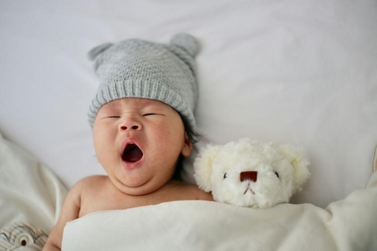 A baby yawns while tucked into bed with a teddy bear