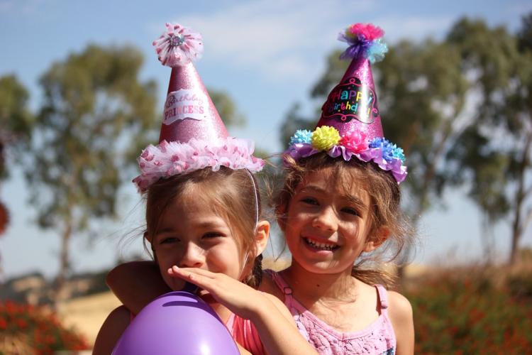 Two little girls in party hats blow up balloon at outdoor children's event