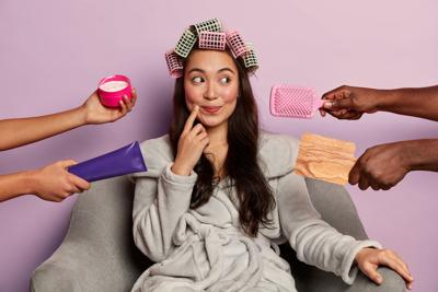 Photo of lovely woman with pensive dreamy expression, wears hair curlers and bathrobe, prepares for