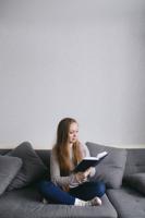 Smart Young Female Sitting On The Couch At Home Reading Book. Beautiful Woman With Long Hair In Mode